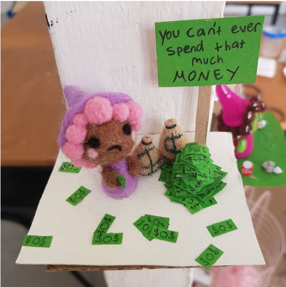 small felt sculpture of an person stood next to a sign that reads "You can't ever spend that much money". surrounded by large quantities of cash and money bags.